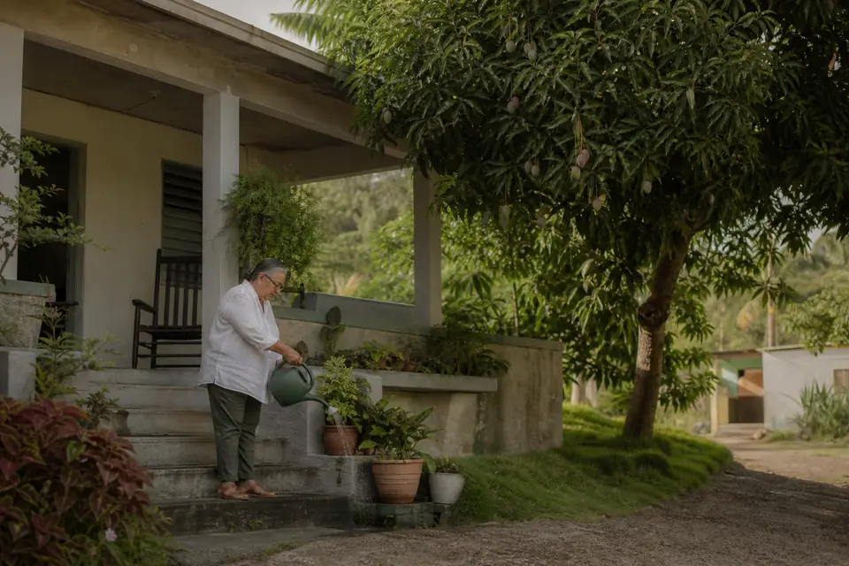 Señora mayor riega plantas frente a su casa familiar en el campo de Utuado